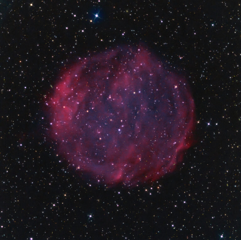 A spherical, faintly glowing pink nebula known as Purgathofer-Weinberger 1 floats against a dark star-filled background. The nebula’s diffuse, circular shape appears semi-transparent, with a mottled texture of filamentary gas and scattered stars shining through and around it. The outer edges glow slightly brighter, forming a faint shell-like structure. Countless distant stars twinkle across the image.