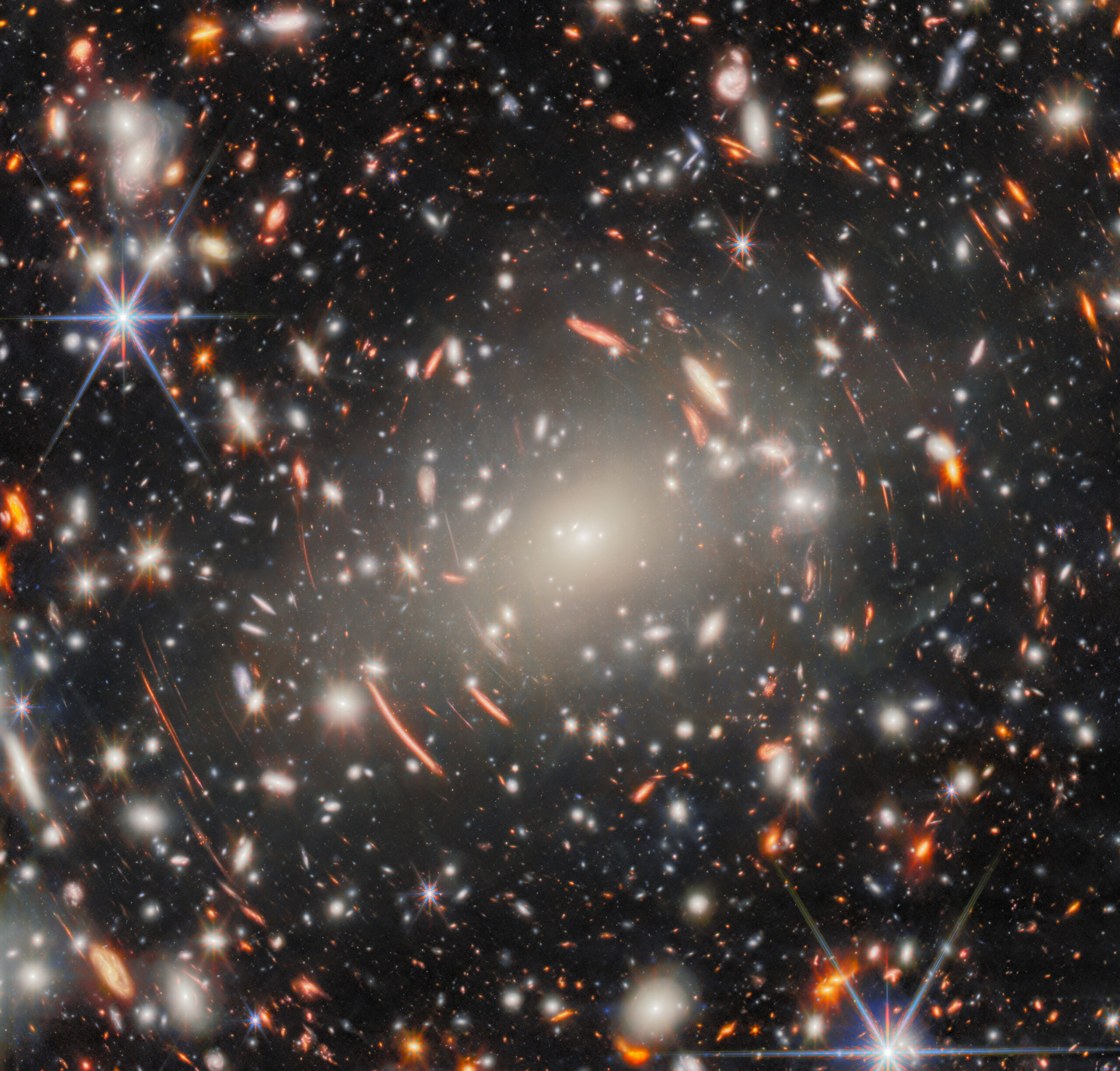 A breathtaking cosmic field filled with bright stars and galaxies, showcasing arcs and streaks of light caused by gravitational lensing. A variety of colors, from red and orange glows to bright white spots, create a mesmerizing, warped effect around a massive galaxy cluster.